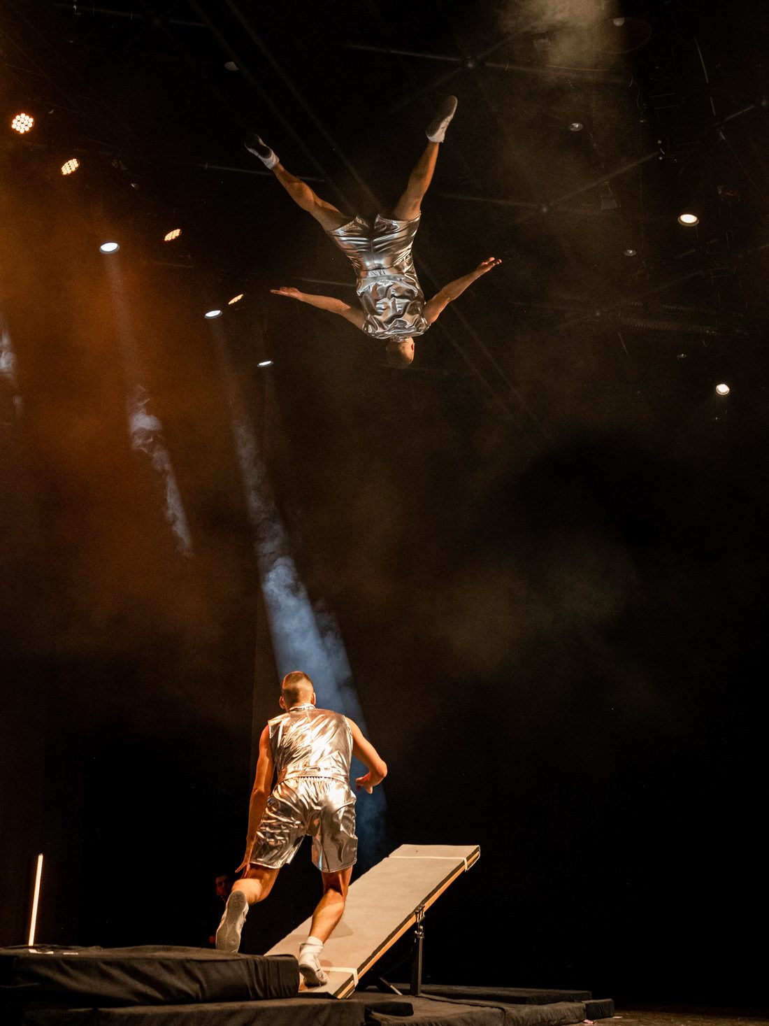 Ein Artist fliegt kopfüber durch den Scheinwerferlichtkegel, ein zweiter läuft auf das Teeterboard zu